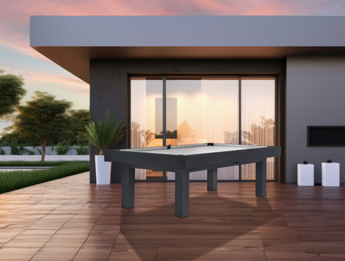 Modern black outdoor pool table with gray playing surface placed on a wooden patio in front of a contemporary house, with large glass doors, greenery, and a sunset sky in the background.