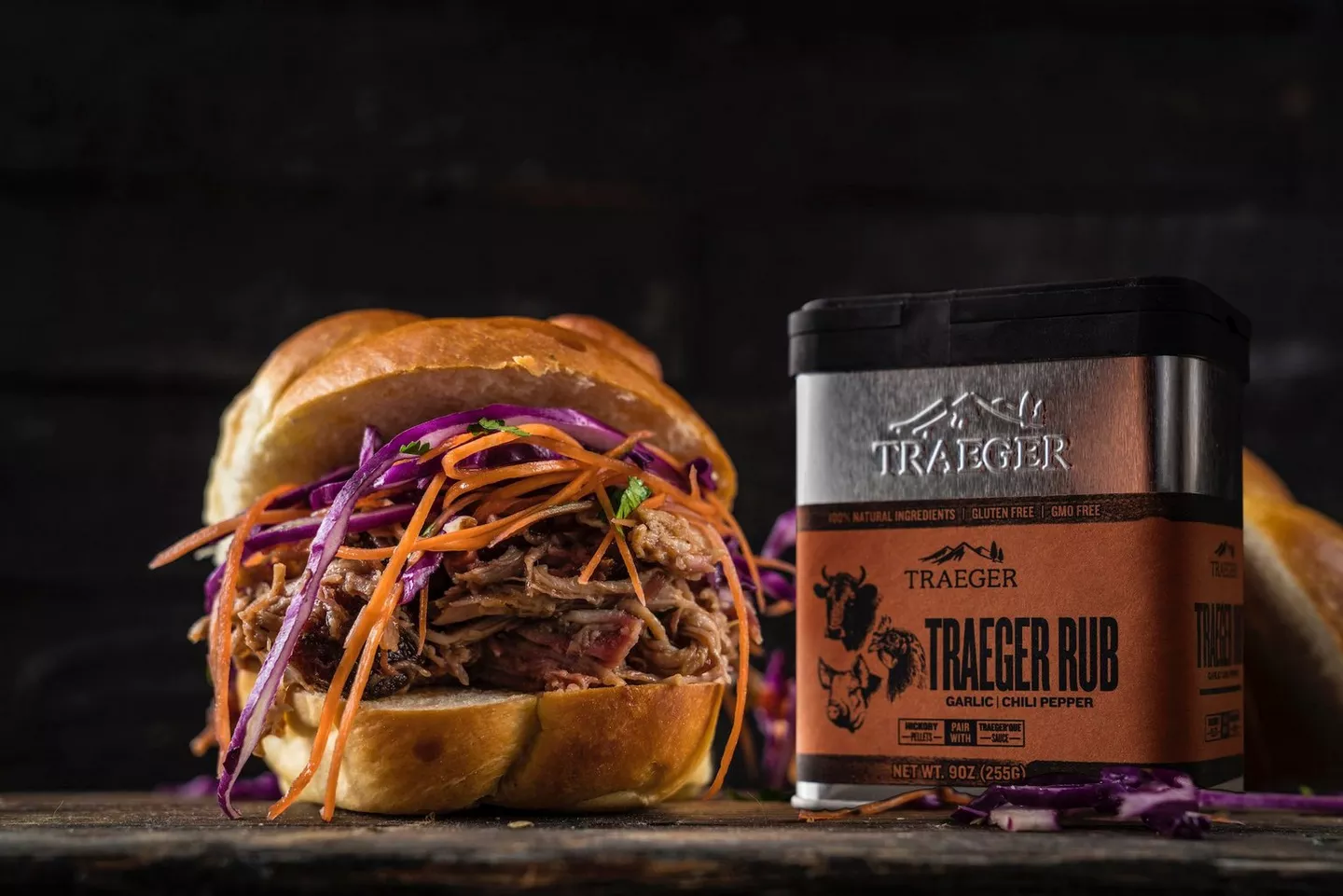 Traeger Rub tin staged beside a pulled-pork sandwich with slaw; dark food-forward composition that gives sauces/spices a real meal setting.