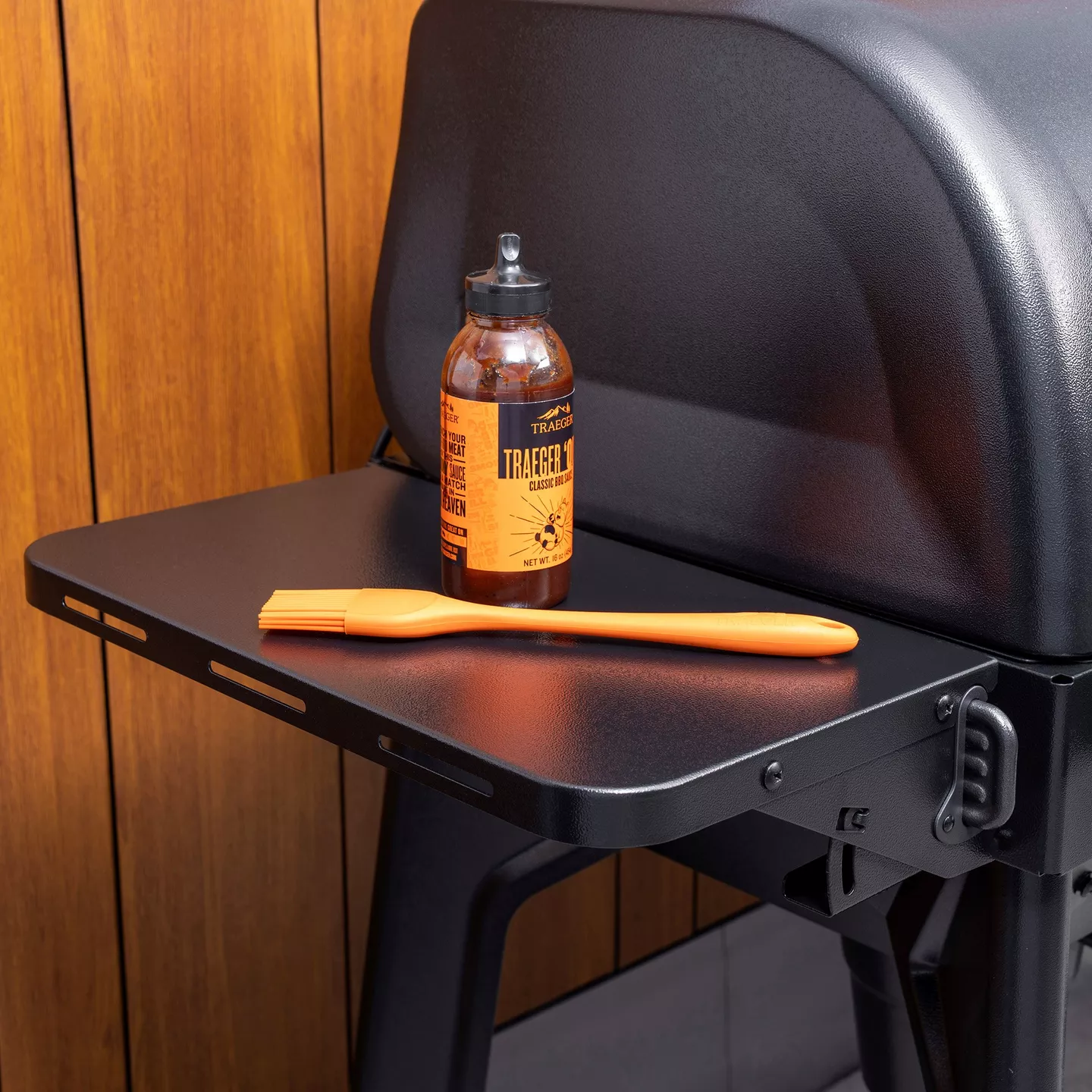 Traeger P.A.L. front shelf attached to a grill, styled with a sauce bottle and basting brush against warm wood paneling; clear in-use accessory context.