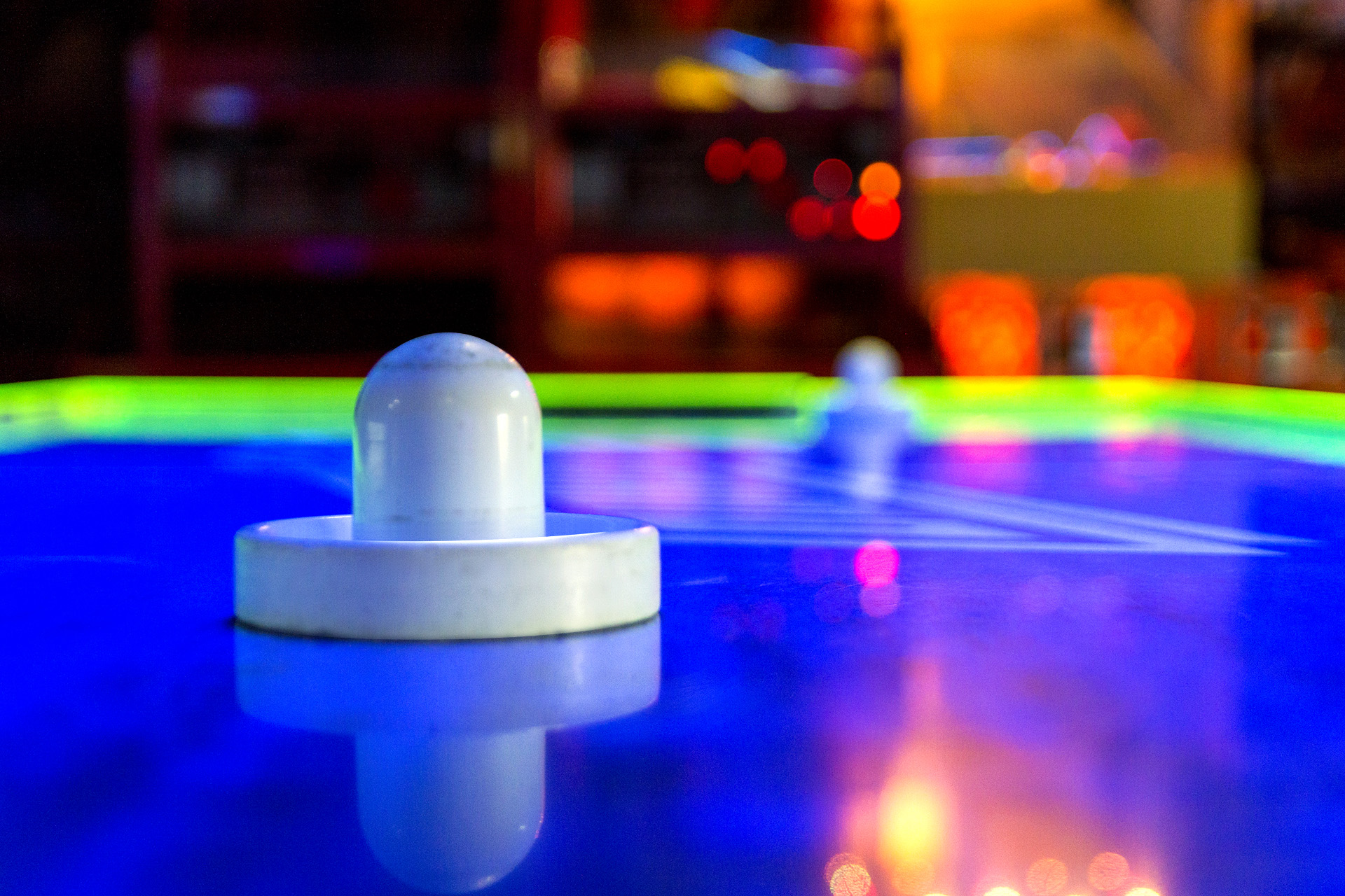 Air hockey pushers on an illuminated playfield with arcade lights in the background