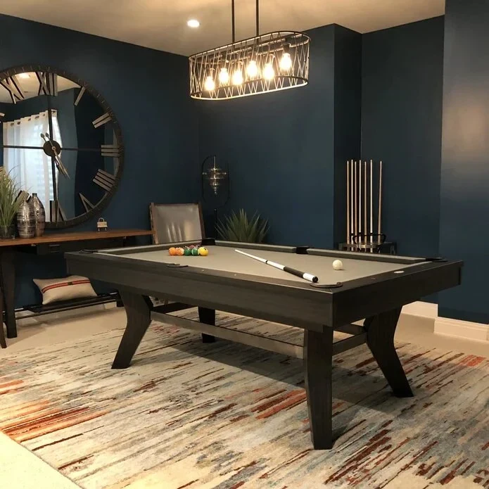 Pool table in a dark blue game room with warm pendant lighting and a large wall clock