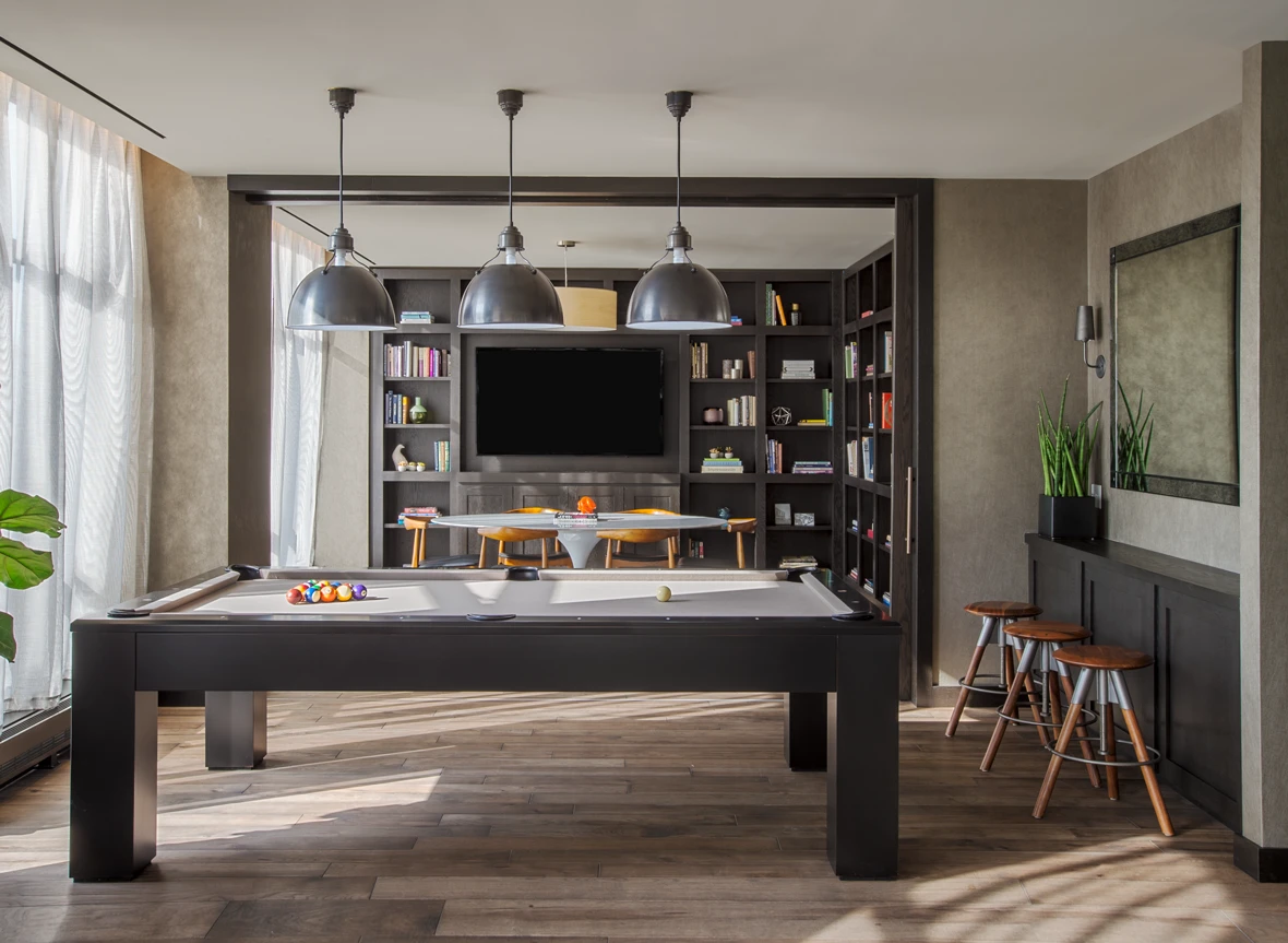 Dark pool table in a sunlit lounge with three metal pendant lights, built-in shelving and wooden stools