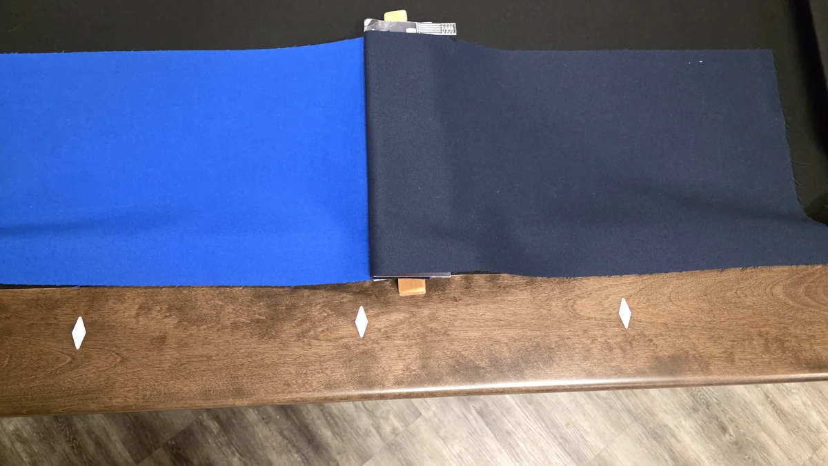Blue and charcoal cloth samples compared on a wood pool table rail