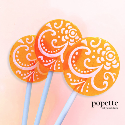 Three round, glossy bright orange hard candy lollipops on top of extra long white paper lollipop sticks. Shown upright in a row with white flower petal designs. All on a light peach background.