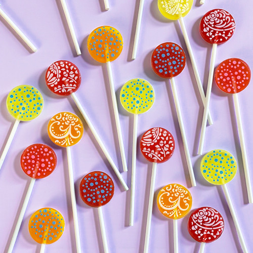 Assortment of lollipops in an overhead photo of several round, glossy hard candy lollipops on top of an extra long white paper lollipop sticks. A bright, multi-colored variety on a light pink/purple background