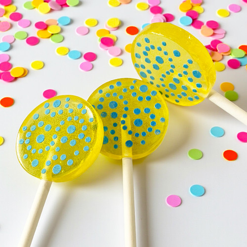 Three bright yellow lollipops with blue polka dots on white sticks. Colorful confetti in pink, yellow, green, and blue is scattered around the lollipops on a white surface.