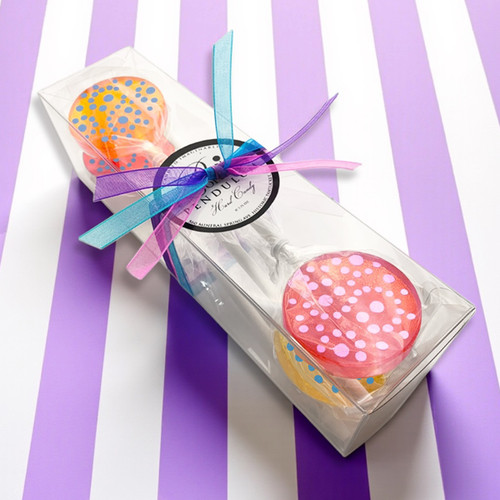 A clear rectangular box containing four round lollipops on sticks. Lollipops are orange, coral pink, yellow and red with frosted dots. The box is tied with a blue, pink and purple ribbon and features a circular label that reads Popette of Pendulum.