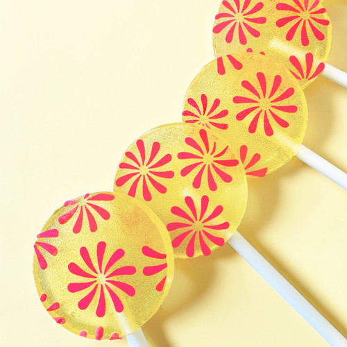 Four bright yellow, round, glossy hard candy Banana Cream Lollipops with extra long white paper lollipop sticks, each with coral pink color pinwheel flavor flowers on a light butter yellow color background.