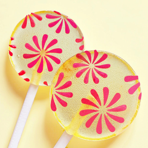 Two bright yellow, round, glossy hard candy Banana Cream Lollipops with extra long white paper lollipop sticks, each with coral pink color pinwheel flavor flowers on a light butter yellow color background.