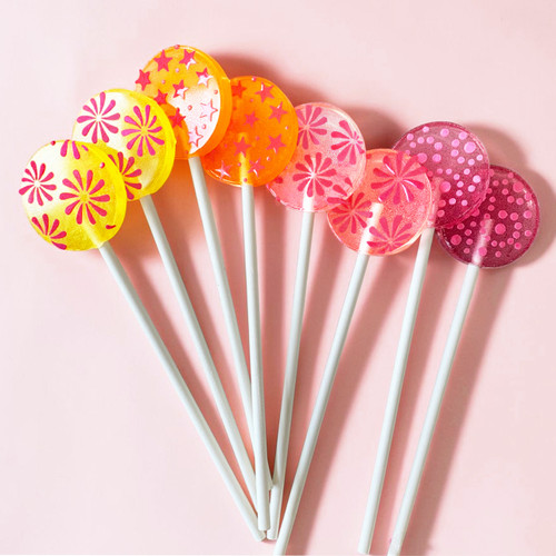 Eight smooth, round, glossy hard candy lollipops on top of extra long white paper lollipop sticks in a fan layout flat lay. Left to Right, our bright yellow Banana Cream lollipop with coral pink flavor flowers. Our bright orange Tropical Punch with coral pink flavor stars. Our light pink Strawberry Banana with coral pink pinwheel flavor flowers and on the top part of the photo is our light purple Berries and Cream with round spiral purple confetti-like flavor dots, all on light pink background.