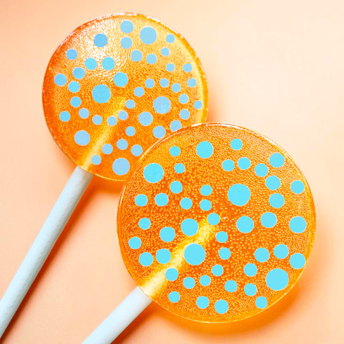 Two bright orange round, glossy hard candy lollipops with extra long white paper lollipop sticks, each with blue polka dot frosting pattern on a light peach background.