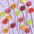 Assortment of lollipops in an overhead photo of several round, glossy hard candy lollipops on top of an extra long white paper lollipop sticks. A bright, multi-colored variety on a light pink/purple background