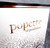 A close-up detail of Lollipop Holder showing white box with the words "popette of pendulum" in a rose-gold colored font. The top edge has a black and rose-gold printed stripe. The bottom of the box features a decorative, floral-like pattern in shades of gray.