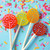 Four smooth, round, glossy brightly colored hard candy lollipops on top of extra long white paper lollipop sticks. In a fan layout with polka dot colored frosting. All on a light teal blue background with multi colored rectangle party confetti.