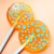 Two round orange lollipops with a translucent, textured surface. Each lollipop features a pattern of light blue circles of varying sizes scattered across the front. The lollipops are attached to smooth, solid white sticks.