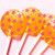 Four bright orange, round, glossy hard candy Tropical Punch Lollipops with extra long white paper lollipop sticks, in a row, each with coral pink color flavor star pattern on a light coral pink background.