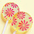 Two bright yellow, round, smooth, glossy hard candy Banana Cream Lollipops with extra long white paper lollipop sticks, each with coral pink color pinwheel flavor flowers on a light butter yellow color background.