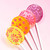 Four smooth, round, glossy hard candy lollipops on top of an extra long white paper lollipop stick. Shown upright in a row, our bright yellow Banana Cream lollipop with coral pink color pinwheel flavor flowers. Our bright orange Tropical Punch with coral pink color flavor stars. Our light pink Strawberry Banana with coral pink pinwheel flavor flowers and on the top part of the photo is our light purple Berries and Cream with round spiral confetti-like flavor dots, all on a light pink background.
