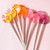 Eight smooth, round, glossy hard candy lollipops on top of extra long white paper lollipop sticks in a fan layout flat lay. Left to Right, our bright yellow Banana Cream lollipop with coral pink flavor flowers. Our bright orange Tropical Punch with coral pink flavor stars. Our light pink Strawberry Banana with coral pink pinwheel flavor flowers and on the top part of the photo is our light purple Berries and Cream with round spiral purple confetti-like flavor dots, all on light pink background.
