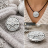 round snap jewelry charm with white enamel and silver snowflake design accented by iridescent AB crystals, shown loose and worn on a brown leather necklace
