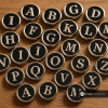 Black and silver SnapAccents snap jewelry charms featuring uppercase alphabet letters A to Z on a wooden surface, customizable interchangeable accessories for bracelets and necklaces.