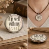 Round SnapAccents snap jewelry charm with inspirational quote "it is well with my soul" in black and beige text, silver metal frame, placed on wooden surface, ideal for interchangeable snap jewelry accessories.