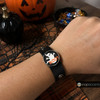 Black snap jewelry bracelet with a round charm featuring a white ghost and orange 'Boo!' text on a black background worn on a wrist with Halloween decorations in the background