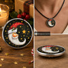 Round snap jewelry charm featuring a crescent moon Santa with a red hat and holly, surrounded by golden stars on a black background, shown loose and worn on a black cord necklace.