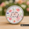 Round SnapAccents snap jewelry charm with watercolor floral design and inspirational text "You Are So Loved," perfect for personalized snap bracelets, necklaces, and accessories, featuring soft pink, purple, and green tones on a wooden surface background.
