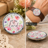 Round SnapAccents snap jewelry charm with watercolor floral design and inspirational text "You Are So Loved," perfect for personalized snap bracelets, necklaces, and accessories, featuring soft pink, purple, and green tones on a wooden surface background.