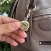 silver snap jewelry charm with a gold heart outline and the word Mom in black script, attached to a brown bag zipper pull