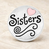 Round silver snap jewelry charm with black script reading Sisters, a pink heart, and swirl details on a beige surface
