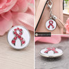snap jewelry charm with a white enamel background and a pink rhinestone ribbon design, shown loose and attached to a beige bag keychain with a tassel