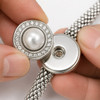 18mm snap jewelry charm with a central white pearl surrounded by a circle of clear rhinestones held near a silver mesh bracelet base