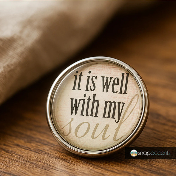 Round SnapAccents snap jewelry charm with inspirational quote "it is well with my soul" in black and beige text, silver metal frame, placed on wooden surface, rustic background, faith-based accessory, interchangeable snap button for bracelets or necklaces.