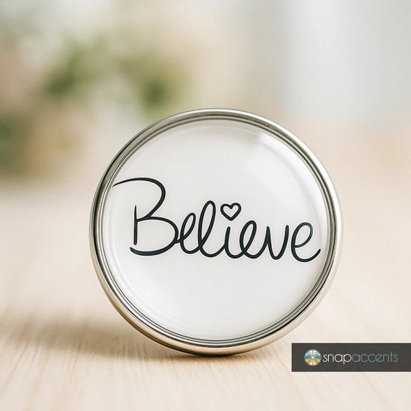 SnapAccents round snap jewelry charm with the word "Believe" in elegant black script and a heart detail, silver metal frame, inspirational accessory for interchangeable snap jewelry, perfect for bracelets, necklaces, and gifts.
