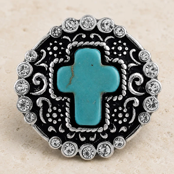 round snap jewelry charm with a turquoise cross center, surrounded by clear rhinestones and silver-toned filigree on a black background