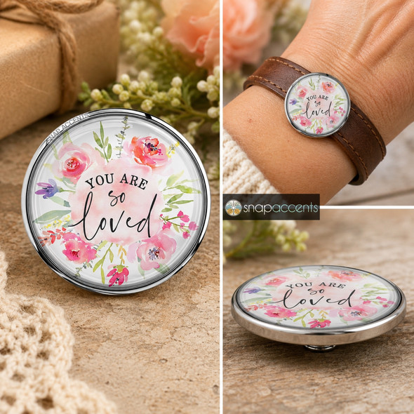 Round SnapAccents snap jewelry charm with watercolor floral design and inspirational text "You Are So Loved," perfect for personalized snap bracelets, necklaces, and accessories, featuring soft pink, purple, and green tones on a wooden surface background.