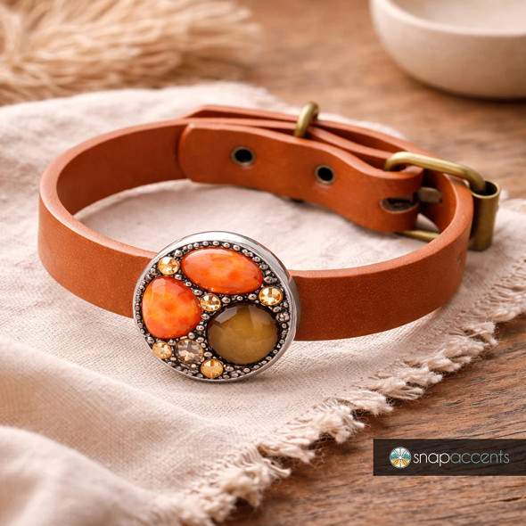 Brown leather adjustable snap bracelet with round decorative snap charm featuring orange and amber gemstones, vintage brass buckle, displayed on beige fabric and wooden surface, SnapAccents jewelry accessory.