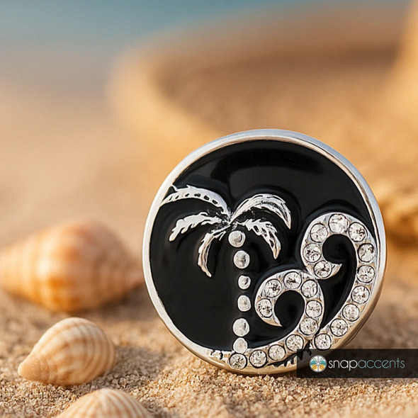 SnapAccents round snap jewelry charm with black enamel background, silver palm tree and wave design embellished with clear rhinestones, beach-themed interchangeable accessory, perfect for bracelets or necklaces, stylish coastal fashion piece on sandy surface with seashells.