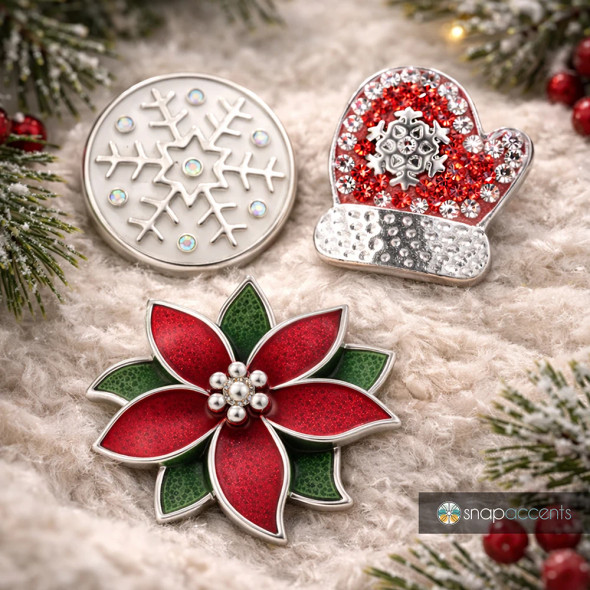 SnapAccents holiday snap jewelry set featuring a red and green poinsettia flower, silver snowflake with iridescent gems, and red mitten with rhinestone snowflake, perfect festive interchangeable snap charms for bracelets or accessories, displayed on cozy beige knit fabric with pine and red berry holiday decor.