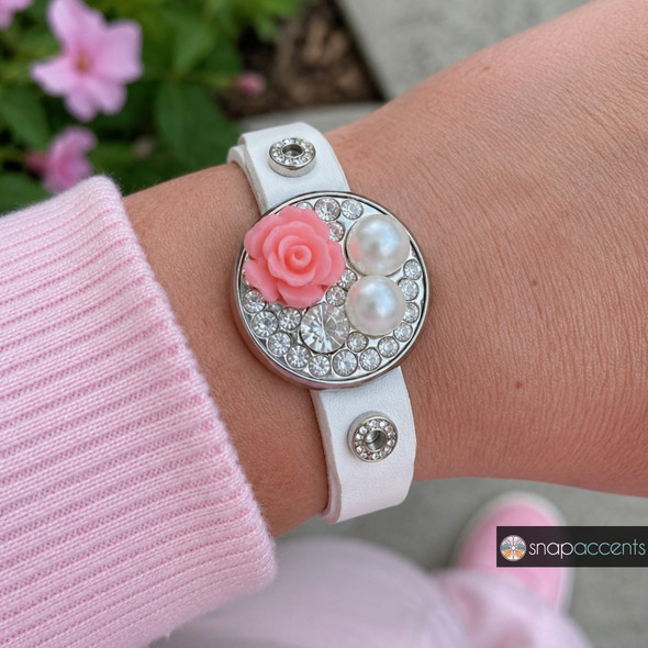 snap jewelry bracelet with a round charm featuring a pink resin rose, two faux pearls, and clear rhinestones on a white leather band