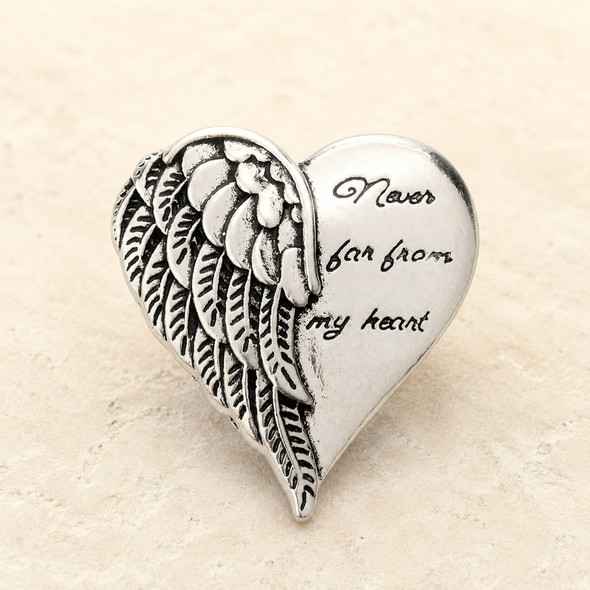 silver-toned snap jewelry charm shaped like a heart with detailed angel wings on the left and the phrase Never far from my heart engraved on the right