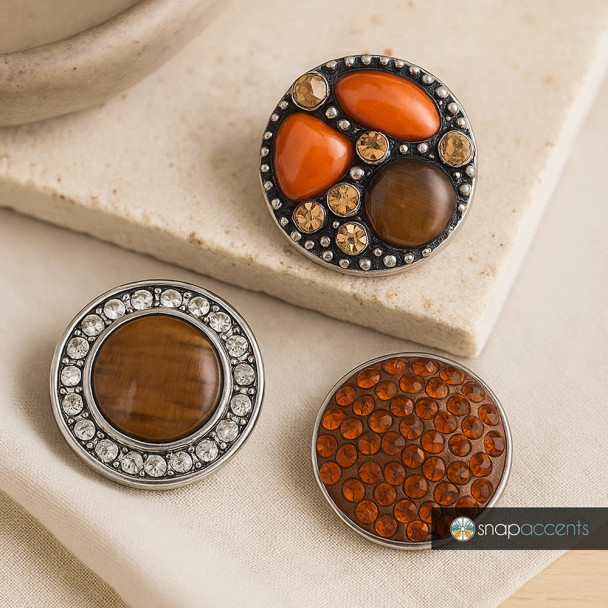 Round SnapAccents snap jewelry buttons featuring tiger's eye stones, orange resin accents, and amber rhinestones in silver-tone settings, perfect for interchangeable charm bracelets and accessories.