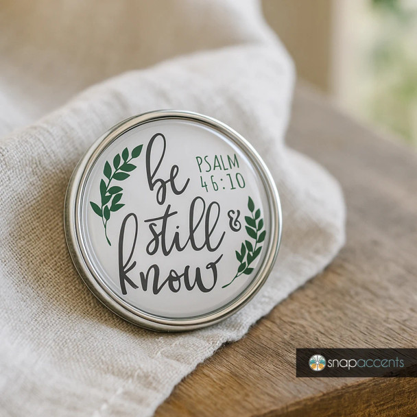 Round SnapAccents snap jewelry charm with "Be Still & Know Psalm 46:10" inspirational scripture text and green leaf design, silver metal frame, displayed on beige fabric and wooden surface, perfect for interchangeable snap jewelry accessories.