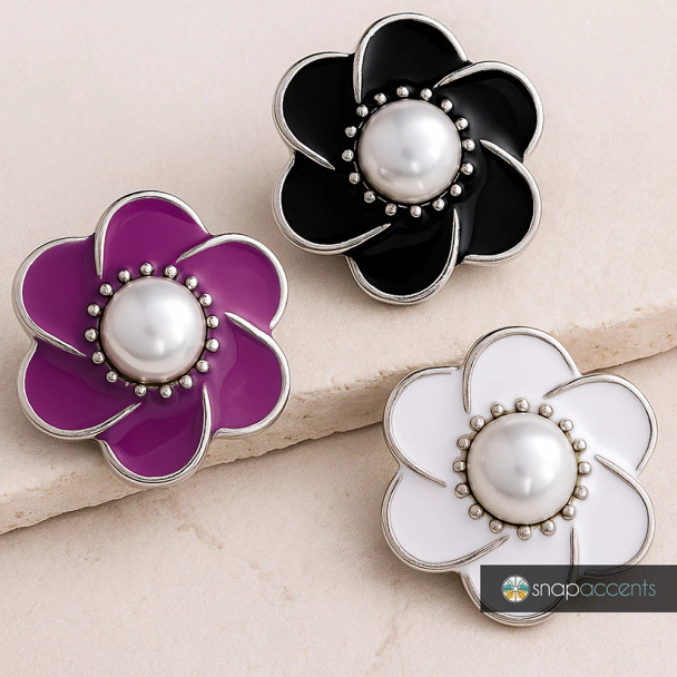 Three SnapAccents snap jewelry flower charms in black, purple, and white enamel with silver edges and central faux pearl accents, displayed on a beige textured surface.