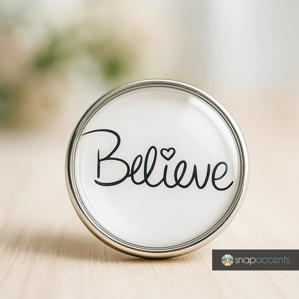 SnapAccents round snap jewelry charm with the word "Believe" in elegant black script and a heart detail, silver metal frame, inspirational accessory for interchangeable snap jewelry, perfect for bracelets, necklaces, and gifts.