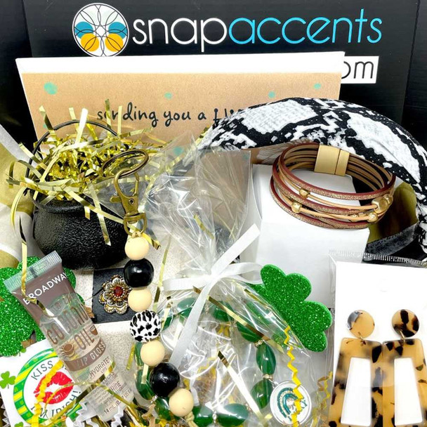 Alt text: SnapAccents St. Patrick’s Day themed jewelry gift set featuring black and cream beaded keychain, green shamrock decorations, tortoiseshell dangle earrings, layered brown and gold bracelets, lip gloss, and snake print headband, all arranged in festive packaging with gold and green accents.