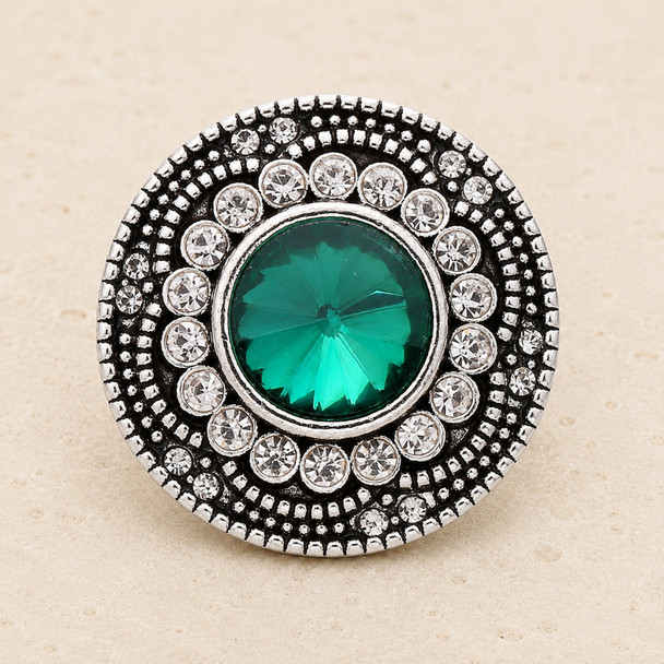 round snap jewelry charm with a faceted green center stone surrounded by two rows of clear rhinestones in a silver-toned setting