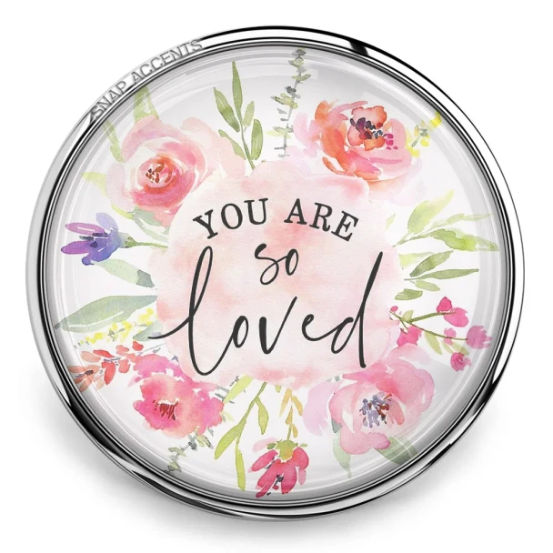 Round SnapAccents snap jewelry charm with watercolor floral design and inspirational text "You Are So Loved," perfect for personalized snap bracelets, necklaces, and accessories, featuring soft pink, purple, and green tones on a wooden surface background.
